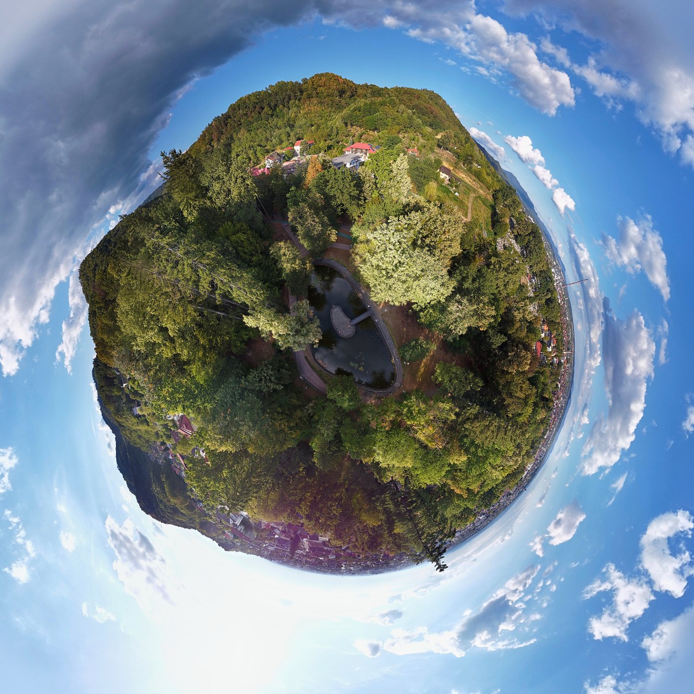 A little planet made from a 360 sphere: a park with a small lake in the middle, trees and houses around it and the sky on the outside